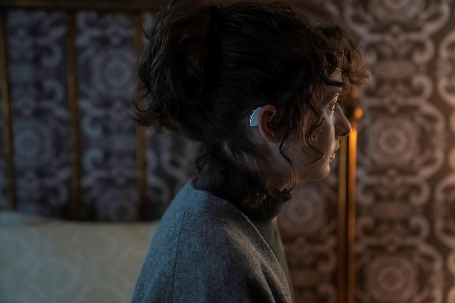 Profil einer Frau mit Hörgerät, grauer Pullover, gemustertem Hintergrund.