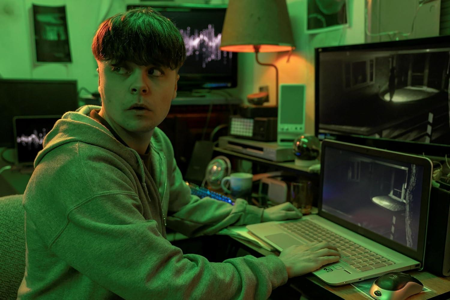 Ein Mann in einem grünen Kapuzenpullover sitzt vor mehreren Monitoren, auf denen Computerbilder zu sehen sind.