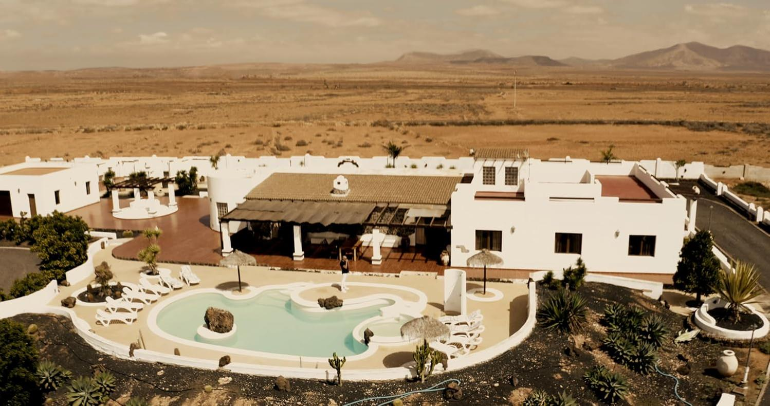 Weiße Gebäude mit Pool und Liegestühlen in trockener Landschaft mit Bergen.