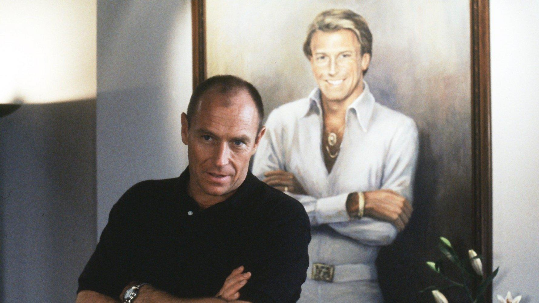 Ein Mann in einem schwarzen Poloshirt steht vor einem Porträt eines anderen Mannes.