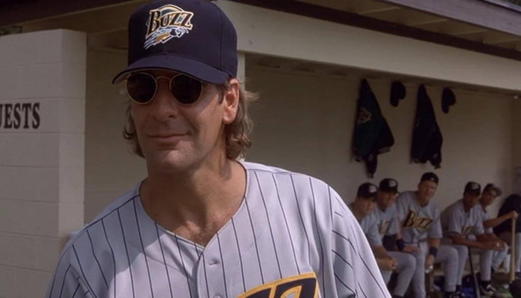 Ein Mann in Baseballkleidung mit Hut und Sonnenbrille steht vor einer Bank mit Spielern.