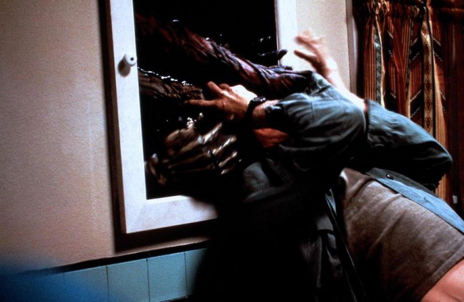 Ein Monsterarm greift einen Mann durch ein Fenster. Dunkle Szene.
