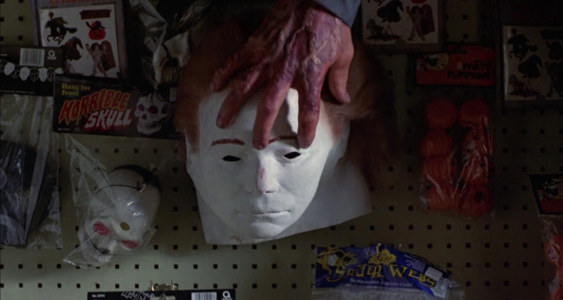Eine Hand auf einer weißen Maske vor Halloween-Dekorationen.