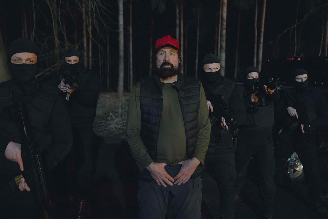 Männer in schwarzer taktischer Ausrüstung mit Waffen stehen in einem dunklen Wald um einen Mann mit roter Kappe.