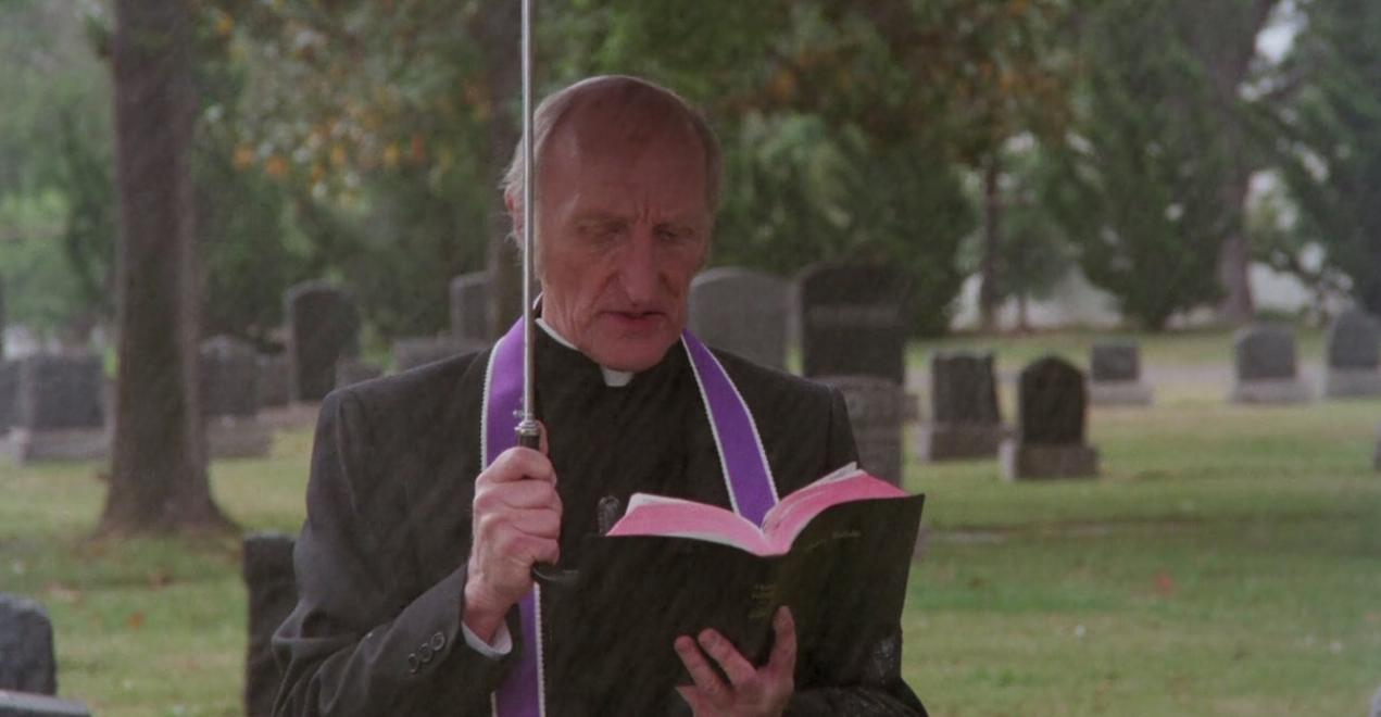 Ein Priester mit lila Schärpe liest bei Regen in einem Friedhof ein Buch.