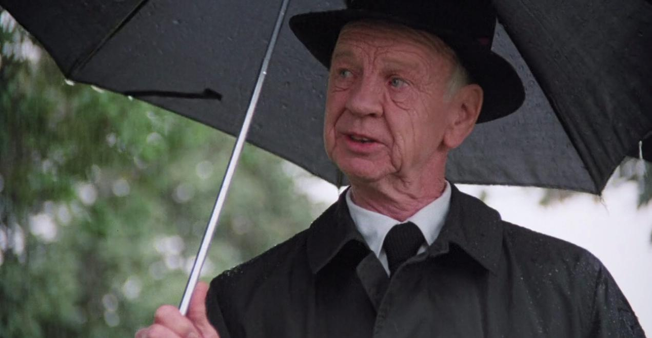 Ein älterer Mann mit Hut und Regenmantel hält einen schwarzen Regenschirm.