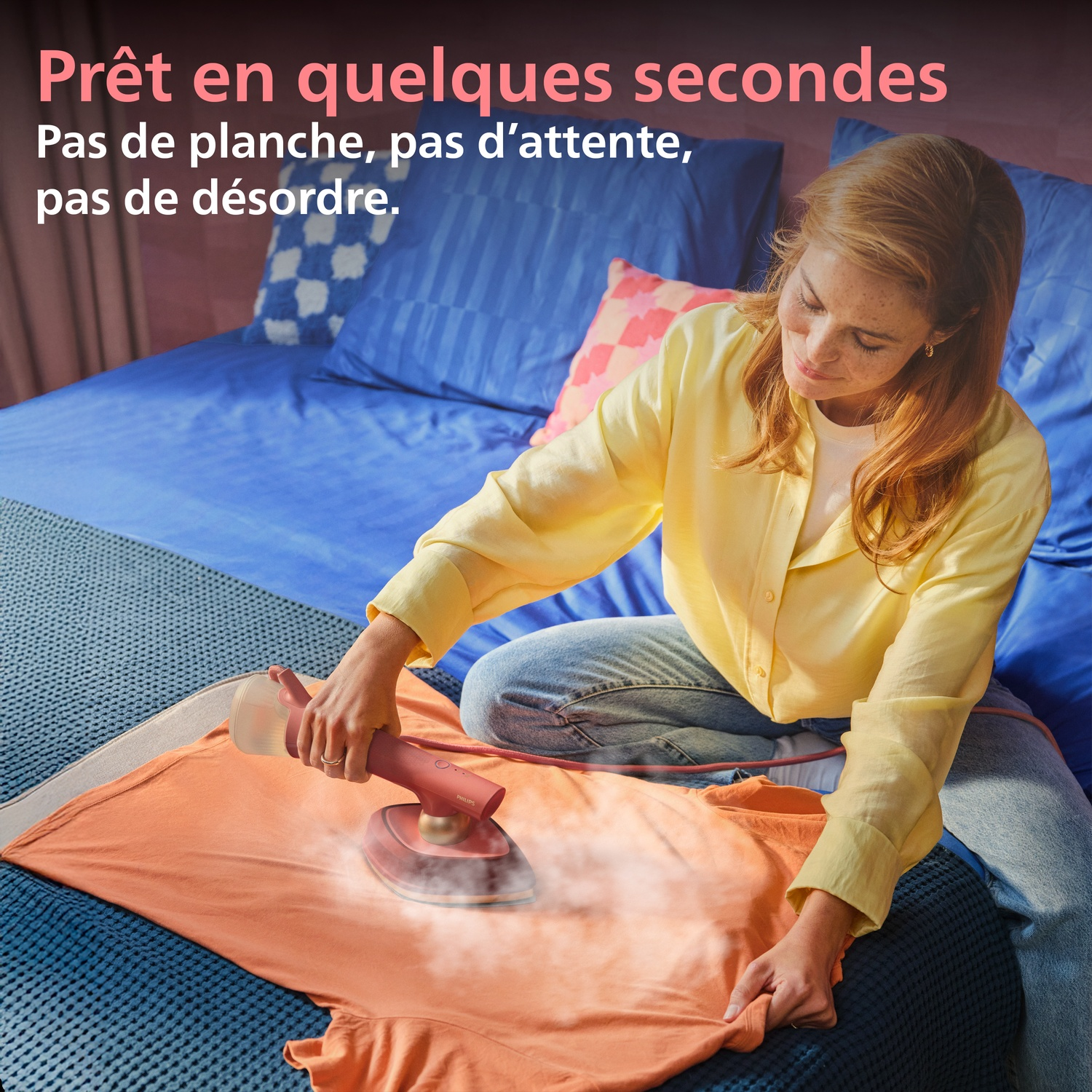 Une femme défroisse un t-shirt orange avec un défroisseur à main sur un lit.