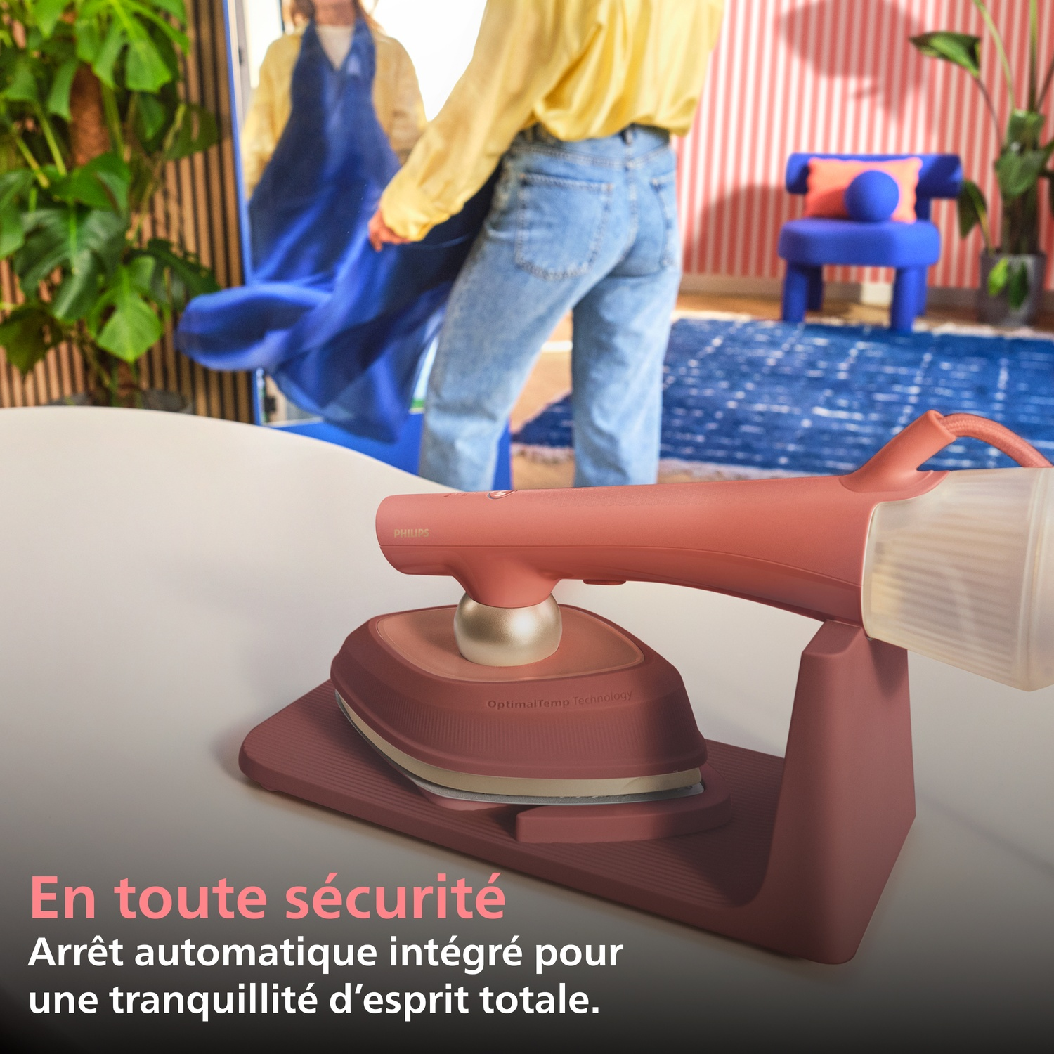 Un fer à vapeur corail repose sur son support. Une personne est visible au fond.