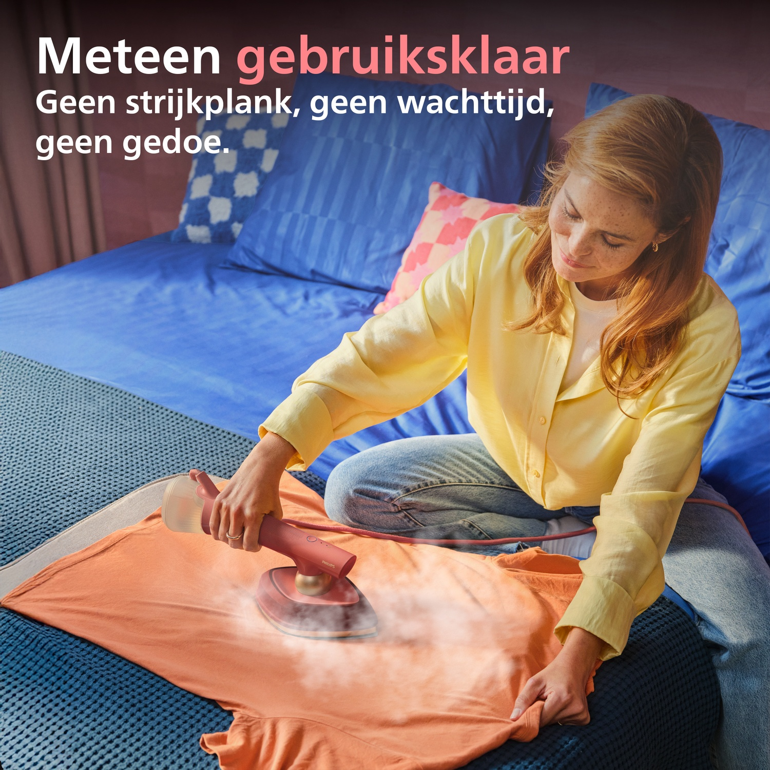 A woman steams an orange t-shirt on a bed with a handheld steamer.