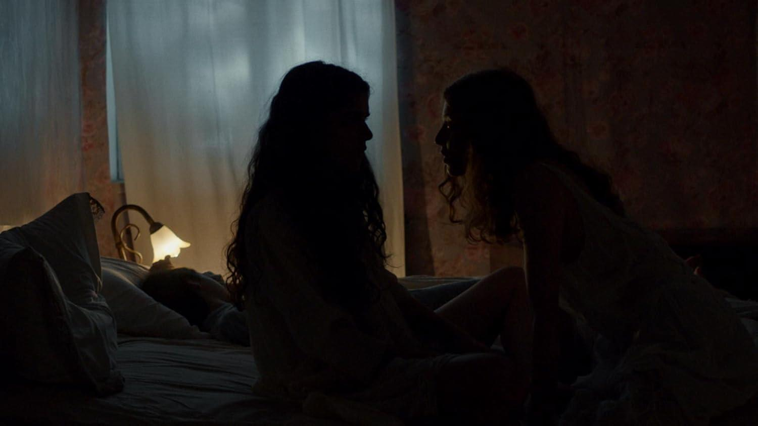 Zwei Frauen sitzen auf einem Bett, eine dritte liegt daneben. Ein Nachttischlampenlicht.