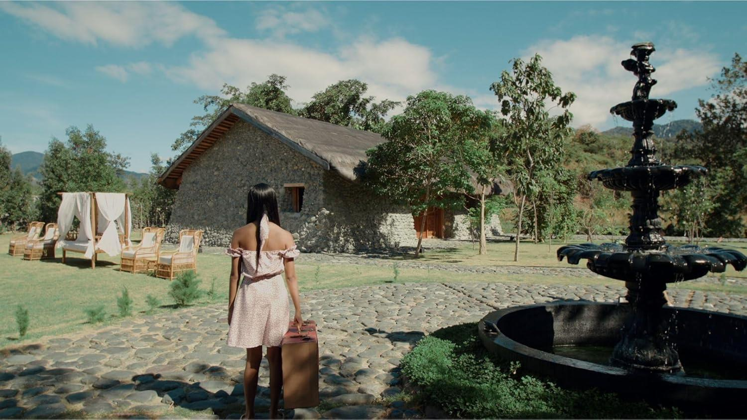 Frau mit Koffer vor Steinhaus, Gartenmöbeln und Brunnen.