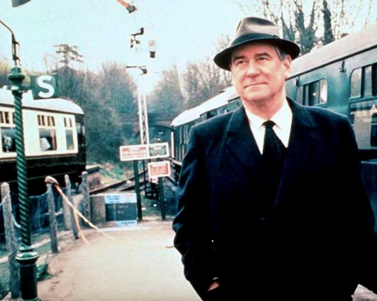 Mann im Anzug und Hut steht auf einem Bahnsteig neben Zügen.