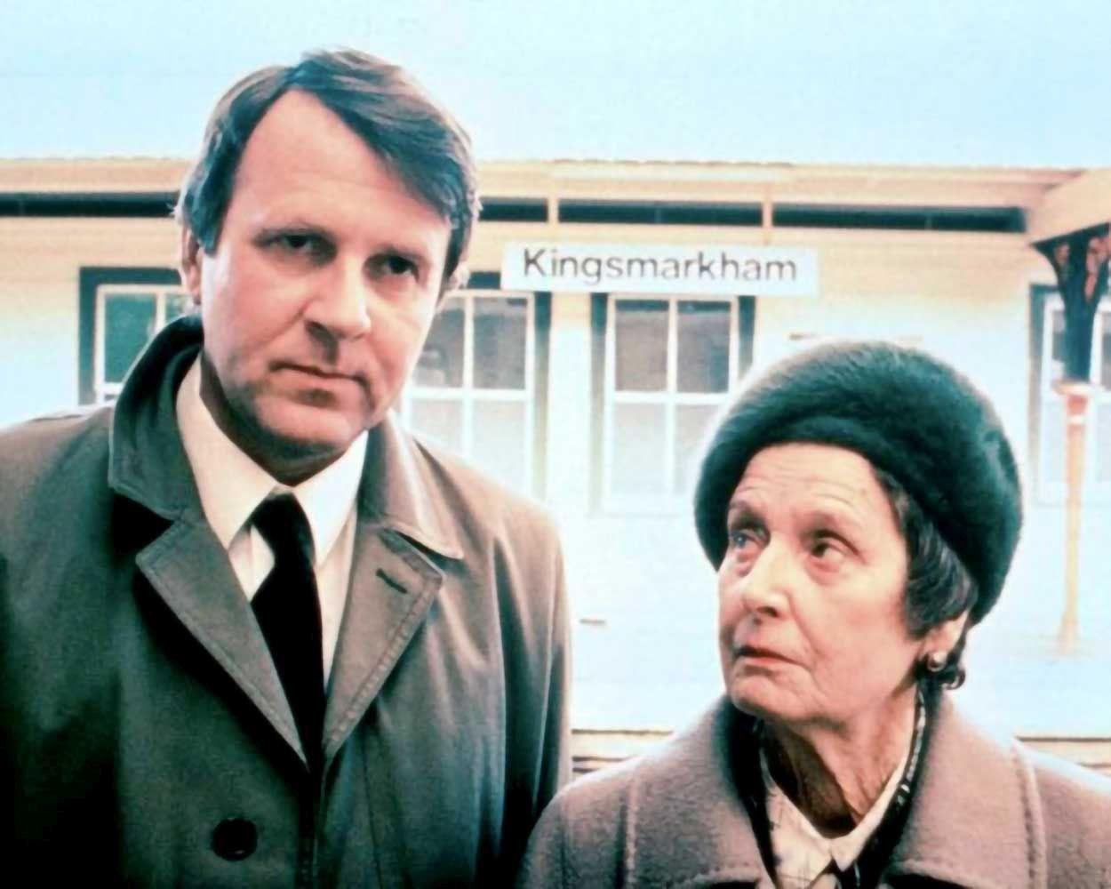 Ein Mann im Trenchcoat und eine Frau mit Hut stehen am Bahnhof Kingsmarkham.