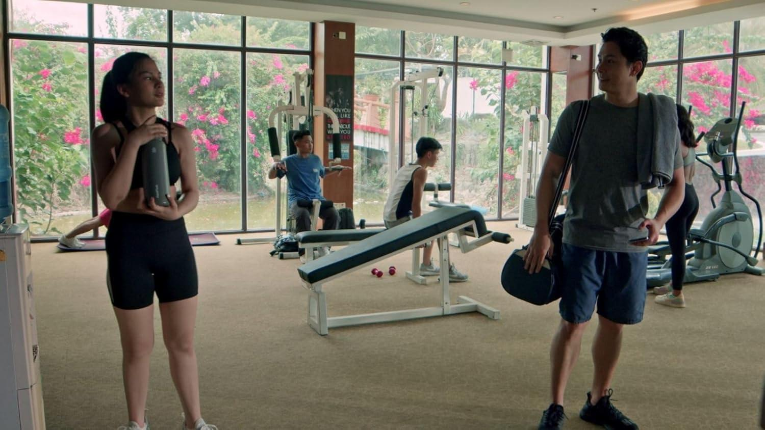 Menschen trainieren in einem Fitnessstudio mit großen Fenstern und Blick auf blühende Pflanzen.