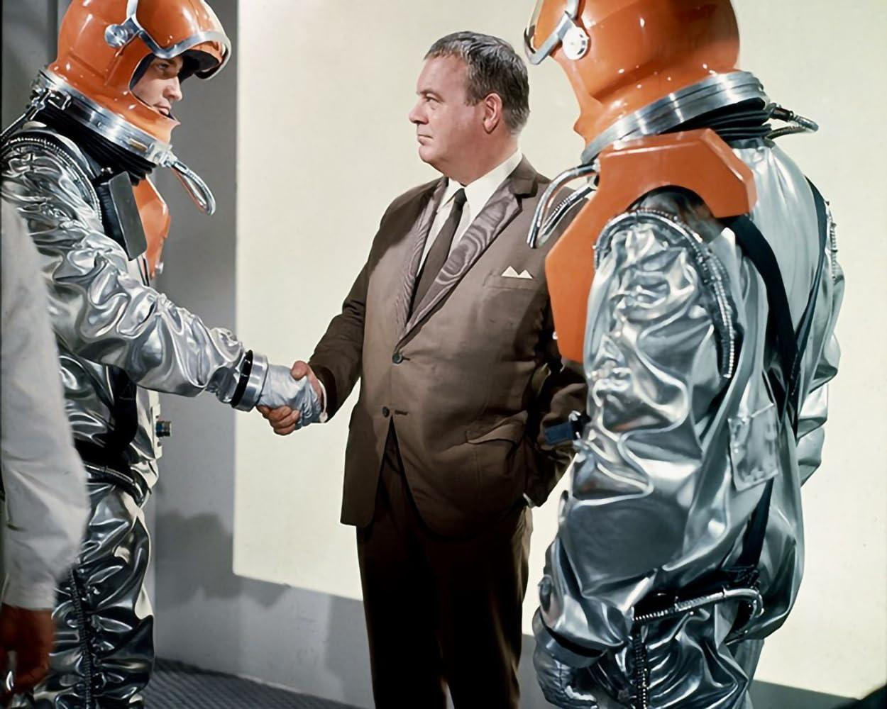 Ein Mann in einem braunen Anzug schüttelt die Hände zweier Astronauten in silbernen Anzügen mit orangefarbenen Helmen.