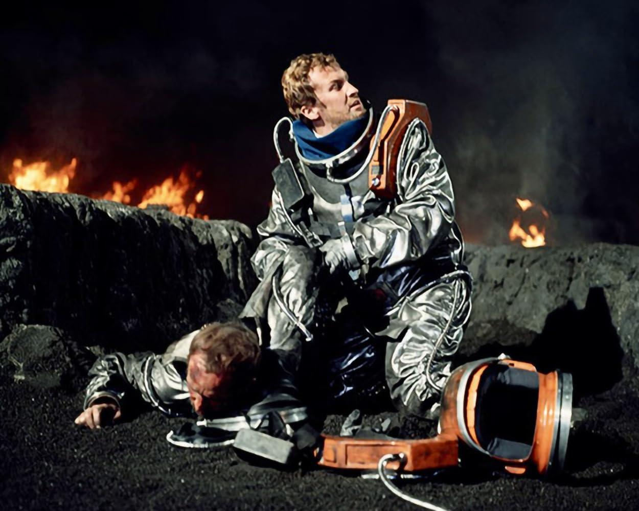 Zwei Astronauten in glänzenden Anzügen, einer kniet, der andere liegt verletzt.