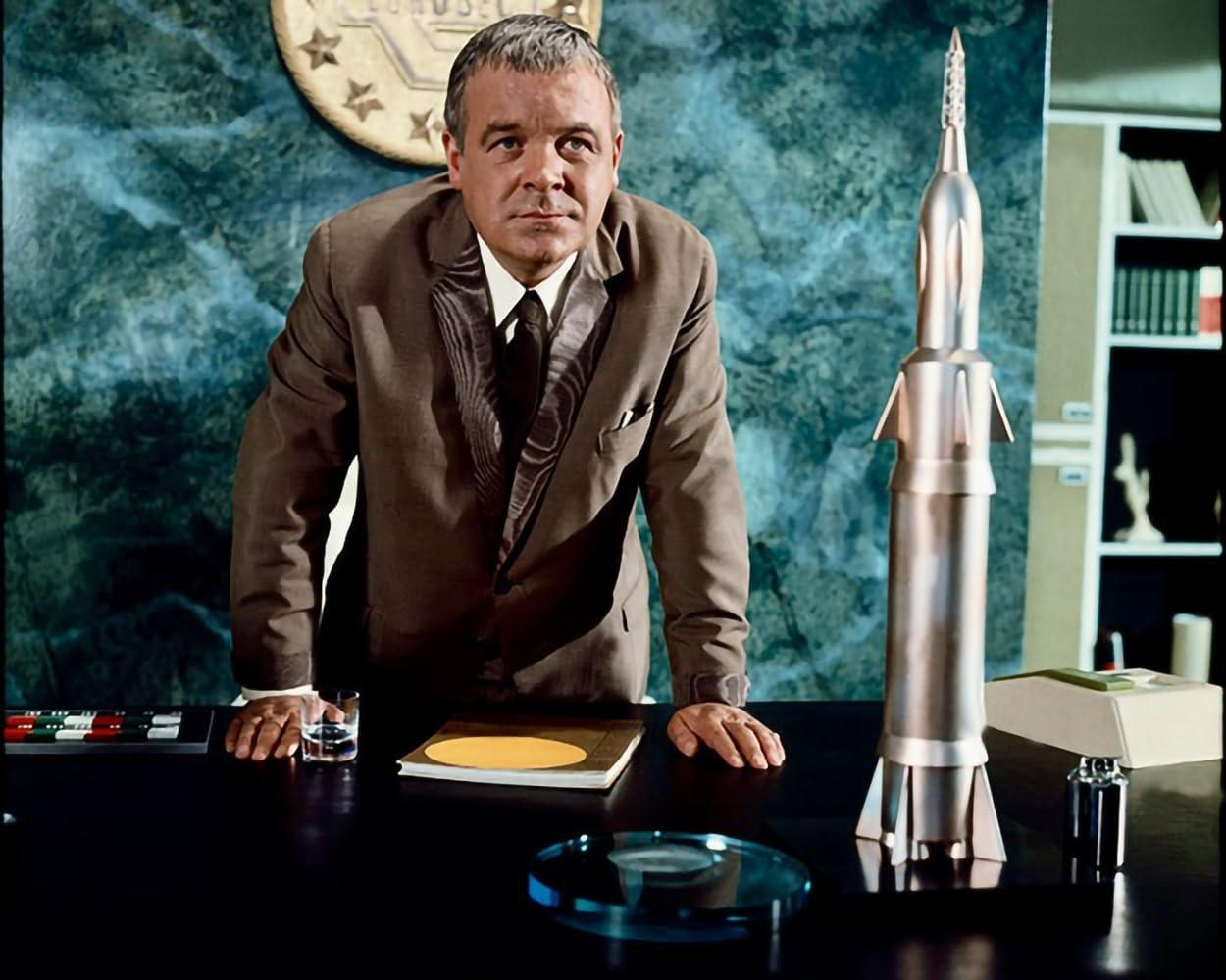Mann im braunen Anzug steht hinter einem Schreibtisch mit Raketenmodell.