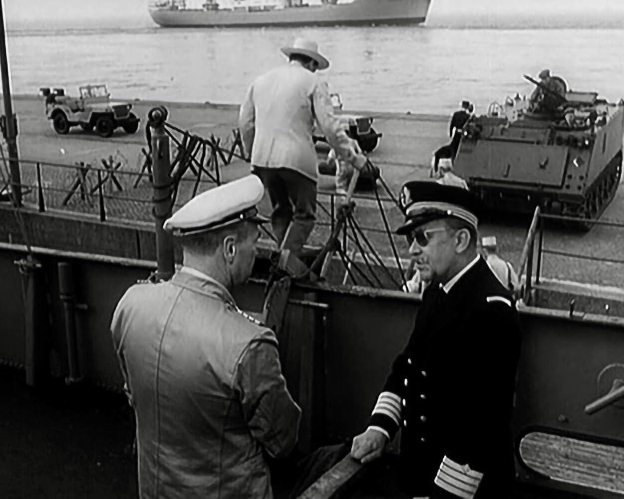 Zwei Männer in Uniform auf einem Schiff. Im Hintergrund ein Panzer und ein großes Schiff.