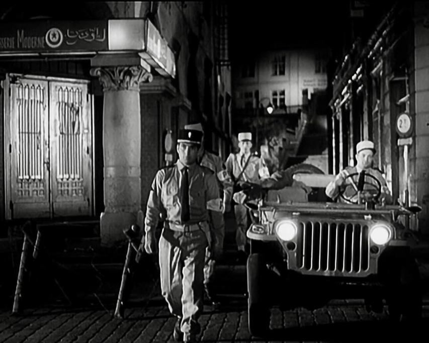 Soldaten in Uniform gehen nachts eine Straße entlang in der Nähe eines Jeeps. Ein Gebäude mit Schild ist sichtbar.