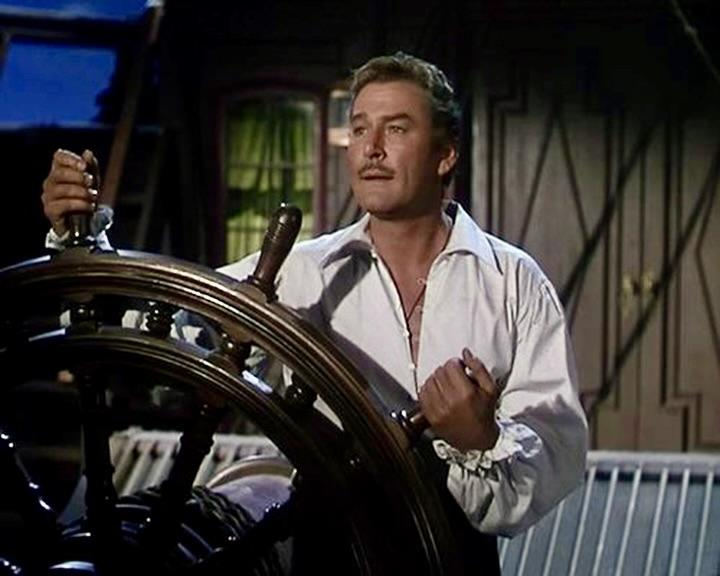 Mann mit Schnurrbart steuert ein Schiff mit einem hölzernen Steuerrad, trägt ein weißes Hemd.