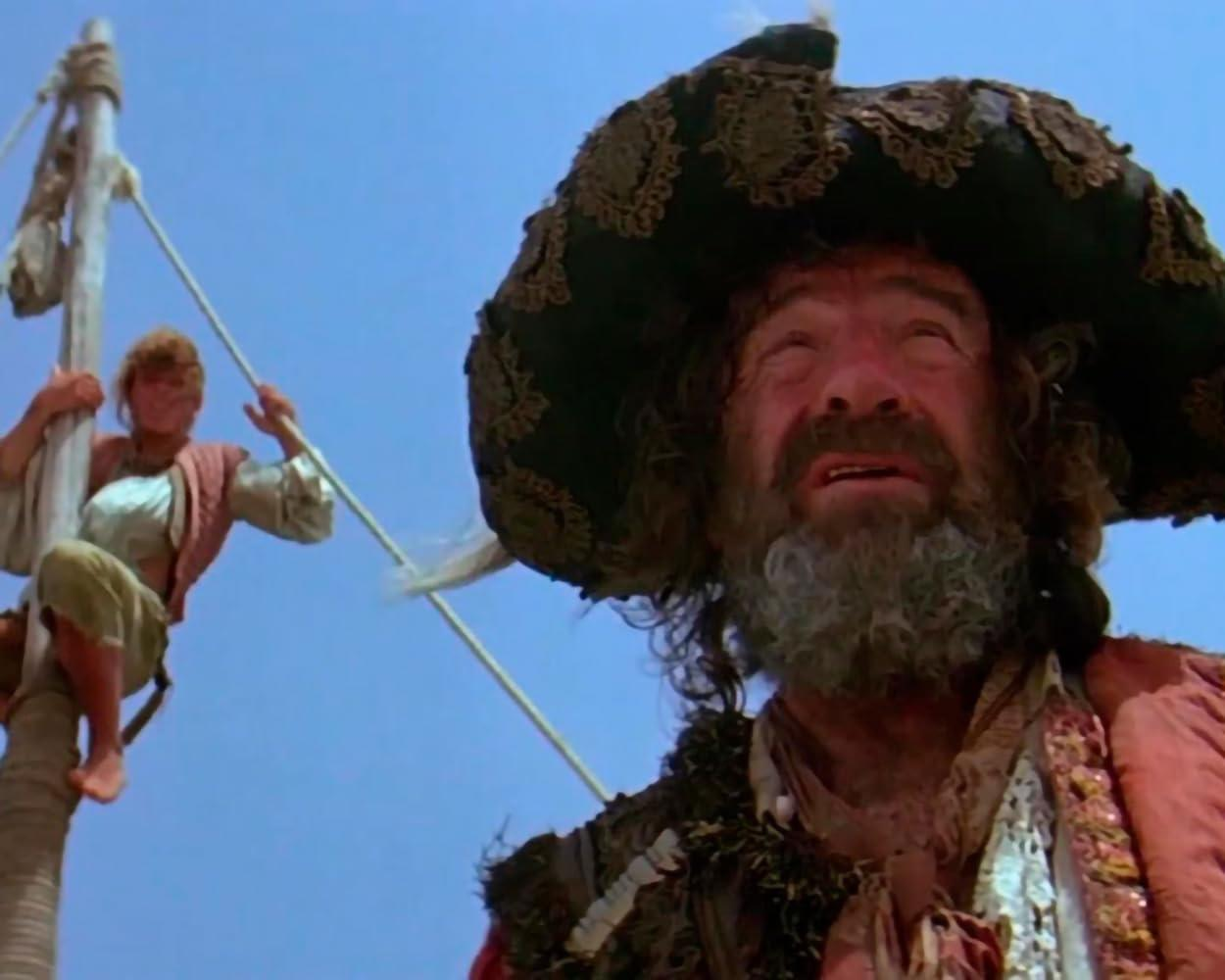 Ein Pirat mit großem Hut und Bart auf einem Schiff. Ein anderer Mann klettert am Mast hoch.