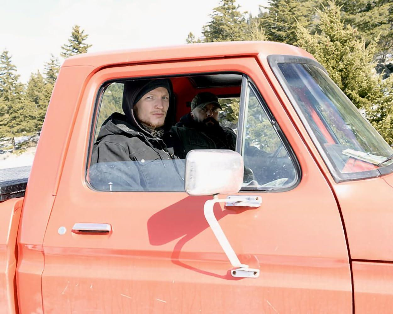 Zwei Männer sitzen in einem orangefarbenen Pick-up-Truck im Freien.