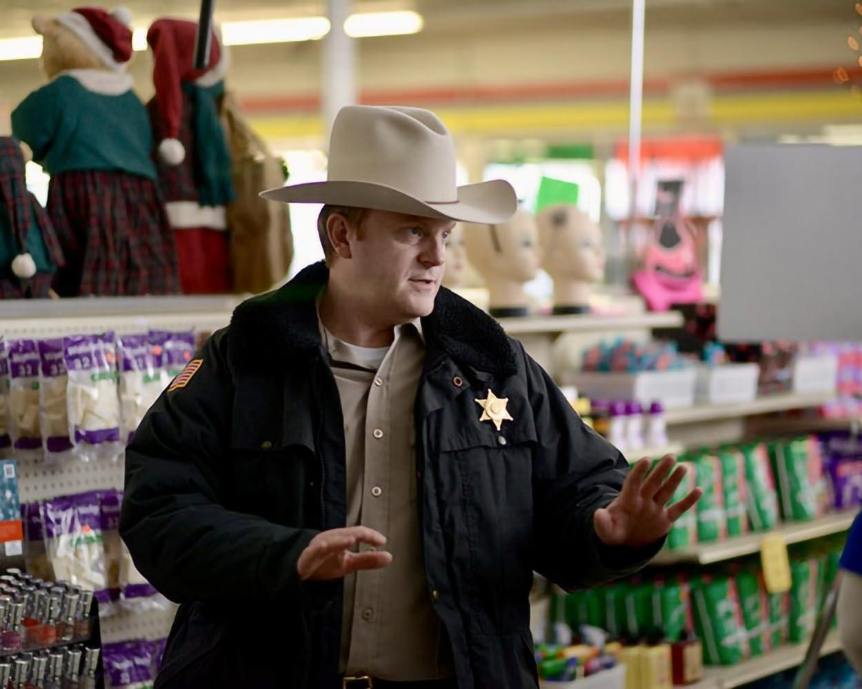 Ein Mann in einem Sheriffhut und schwarzer Jacke steht in einem Geschäft mit Regalen voller Waren.