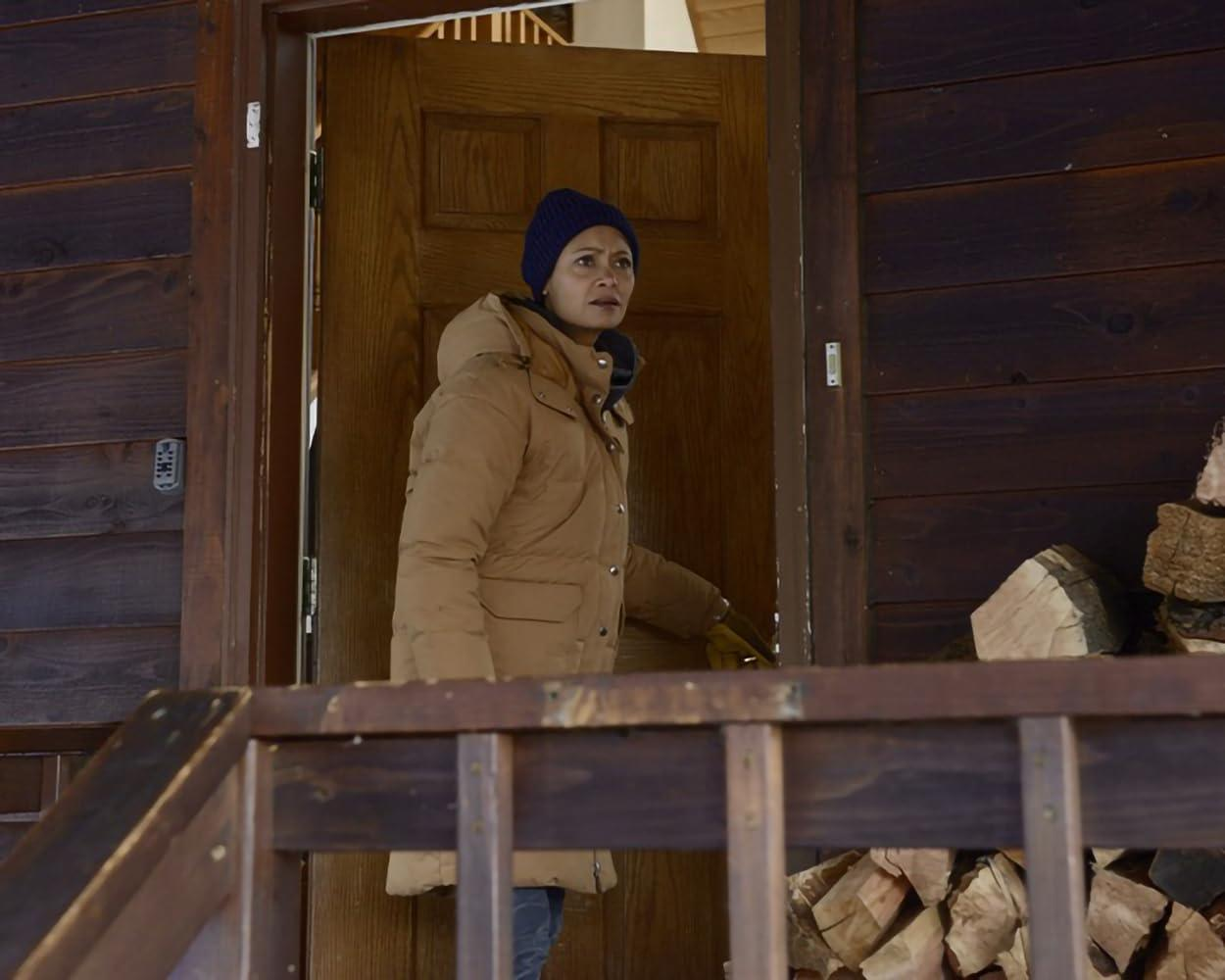 Eine Frau in einem braunen Wintermantel steht im Türrahmen eines Holzhauses.