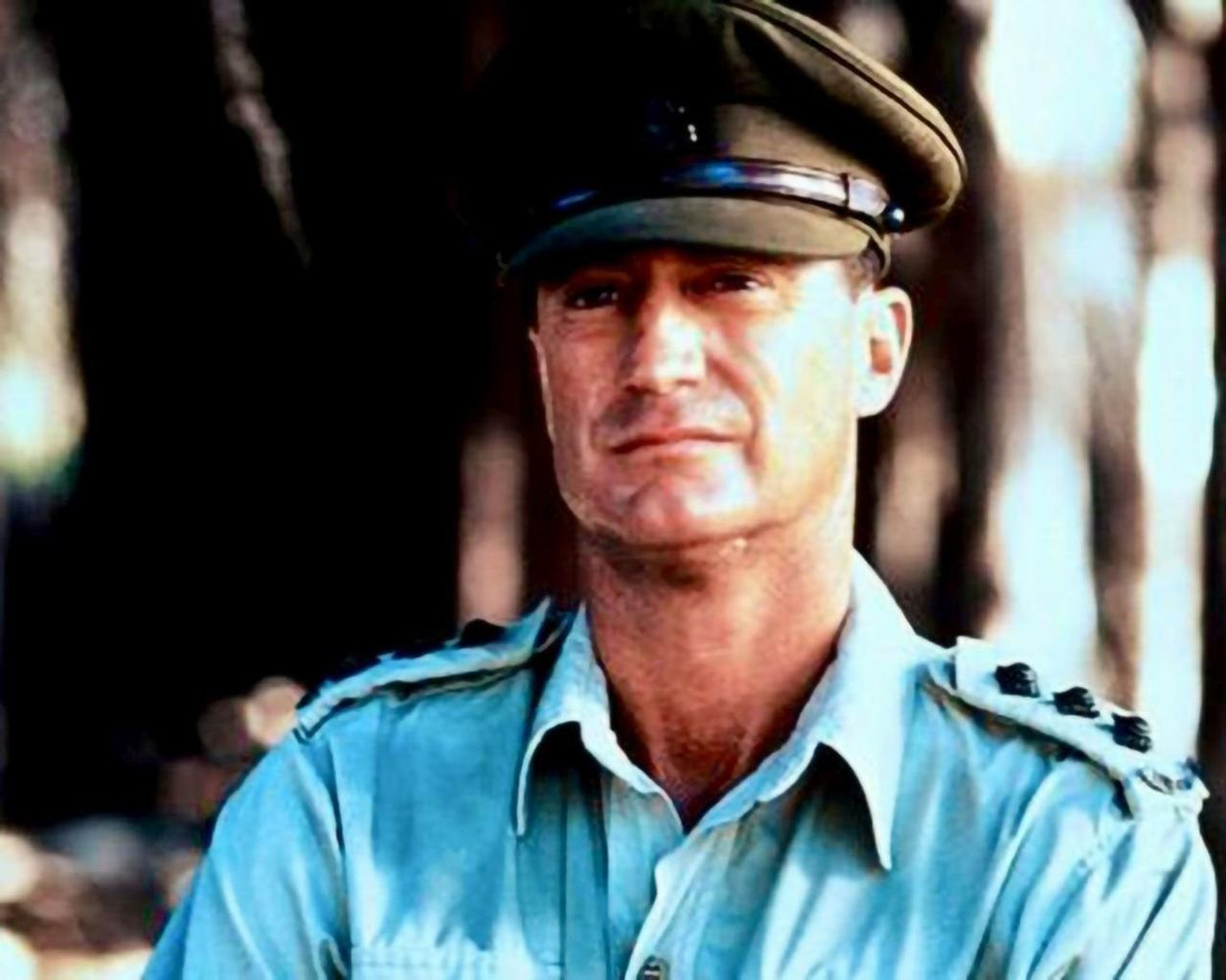 Ein Mann in Uniform mit Hut und blauem Hemd, im Freien mit Bäumen im Hintergrund.