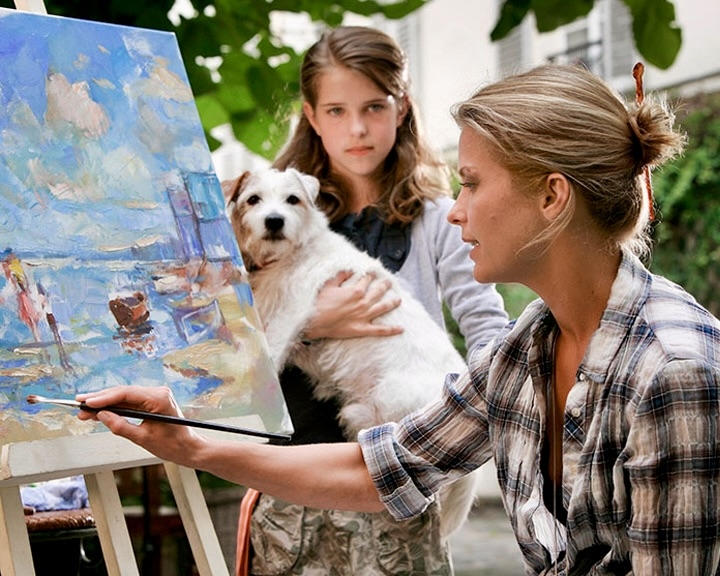 Eine Frau malt ein Bild im Freien. Ein Mädchen hält einen Hund.
