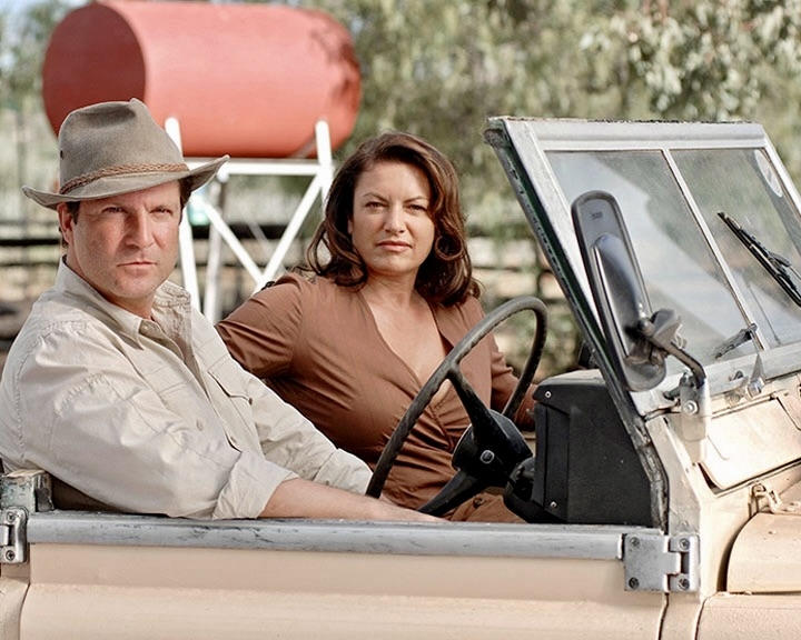 Mann mit Hut und Frau sitzen in einem Auto vor einem roten Tank.