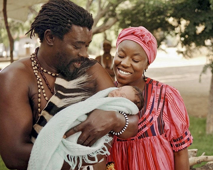 Mann und Frau halten Baby. Mann trägt Halsketten und Fell. Frau trägt rosa Kleid und Turban.