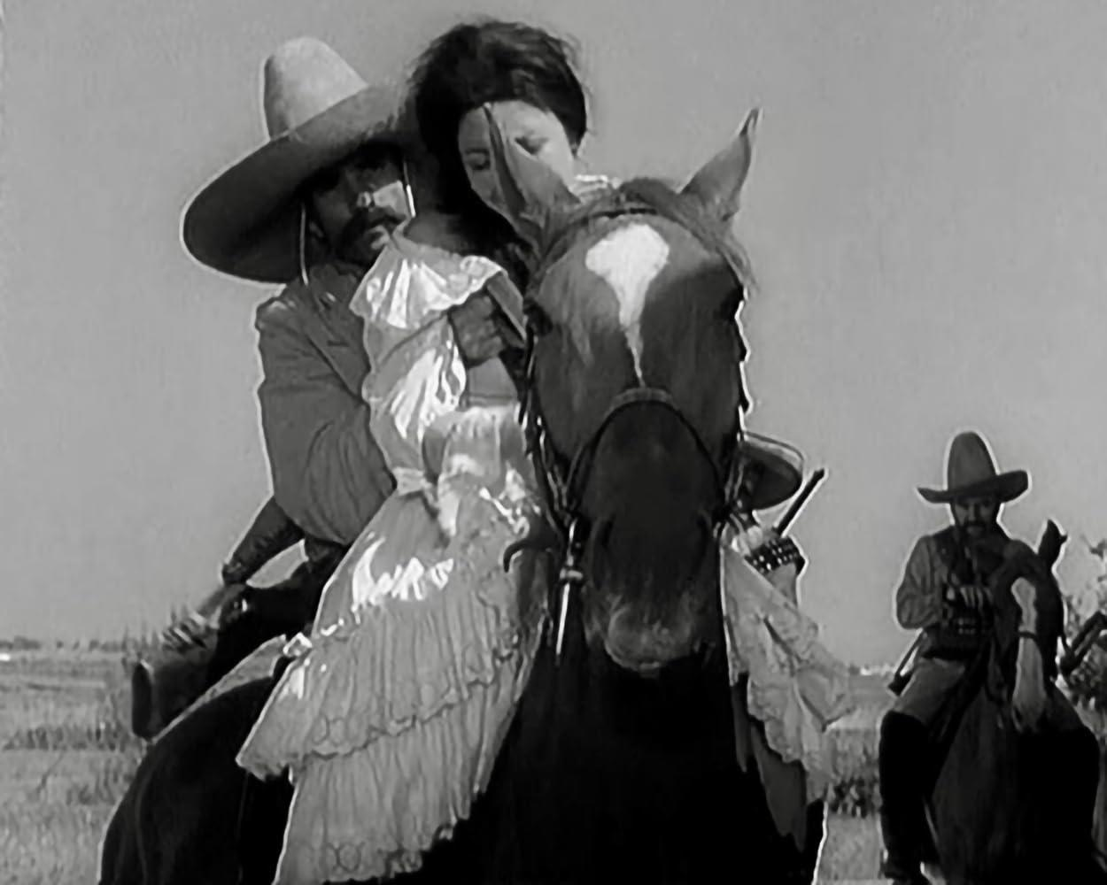 Ein Mann in einem Sombrero reitet mit einer Frau auf einem Pferd. Ein weiterer Reiter ist im Hintergrund.