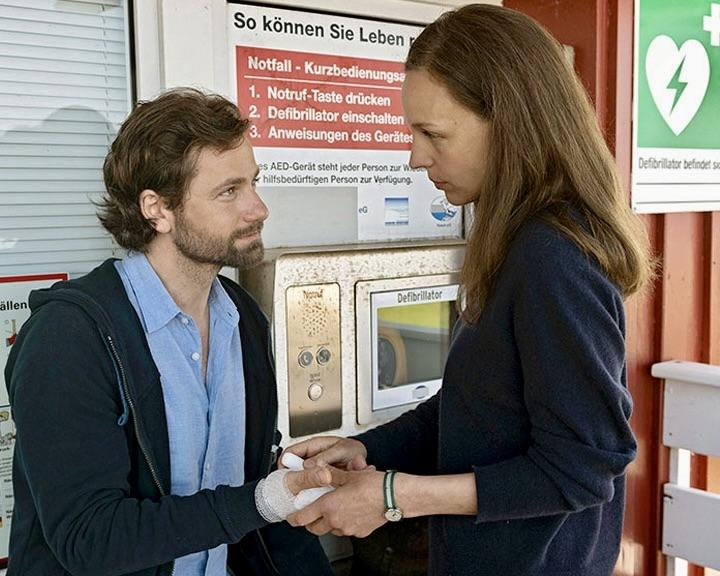 Mann mit verbundenem Arm spricht Frau an, die Defibrillator bedient.