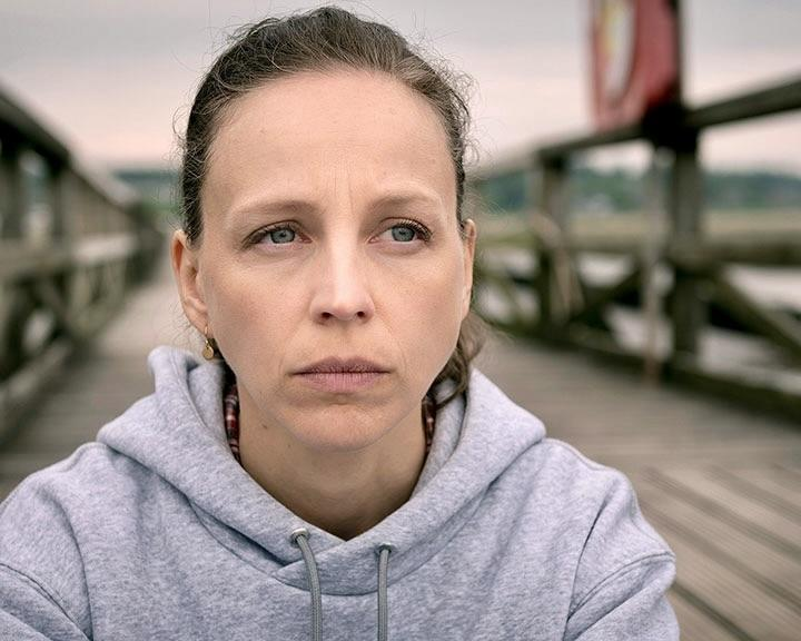 Eine Frau mit grauer Kapuze blickt nachdenklich nach rechts auf einem Holzsteg.