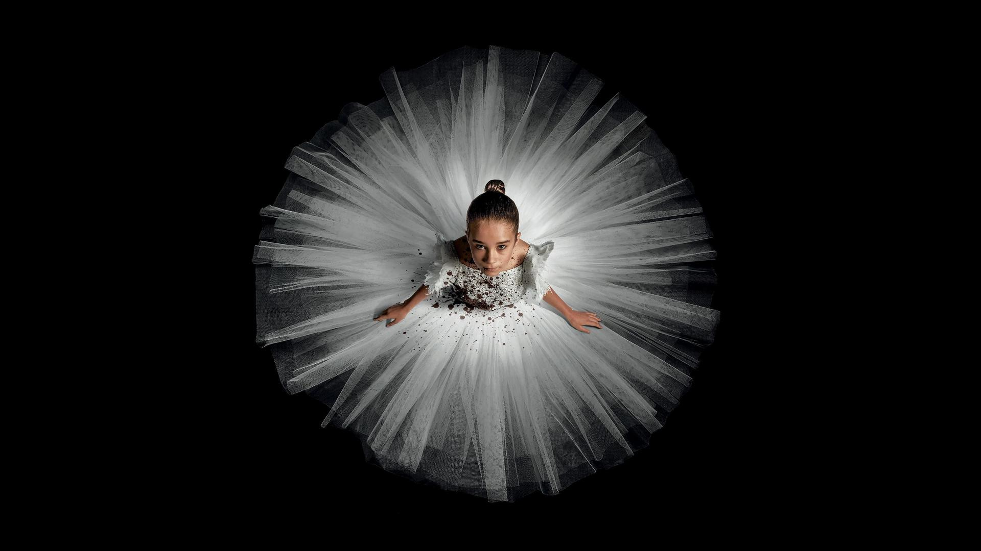 Ein junges Ballettmädchen in einem weißen Tutu mit braunen Spritzern sitzt vor schwarzem Hintergrund.