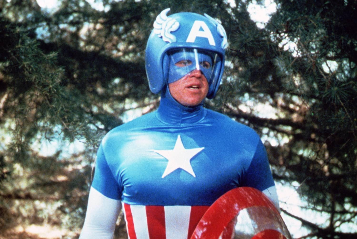 Mann in Captain America Kostüm mit Helm und Schild vor Bäumen.