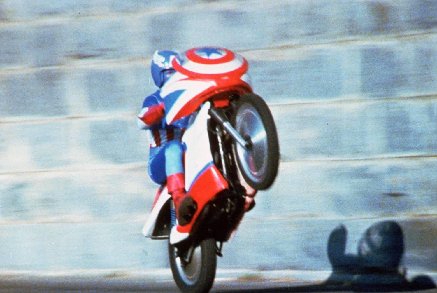 Captain America macht einen Wheelie auf einem Motorrad vor einer blauen Wand.