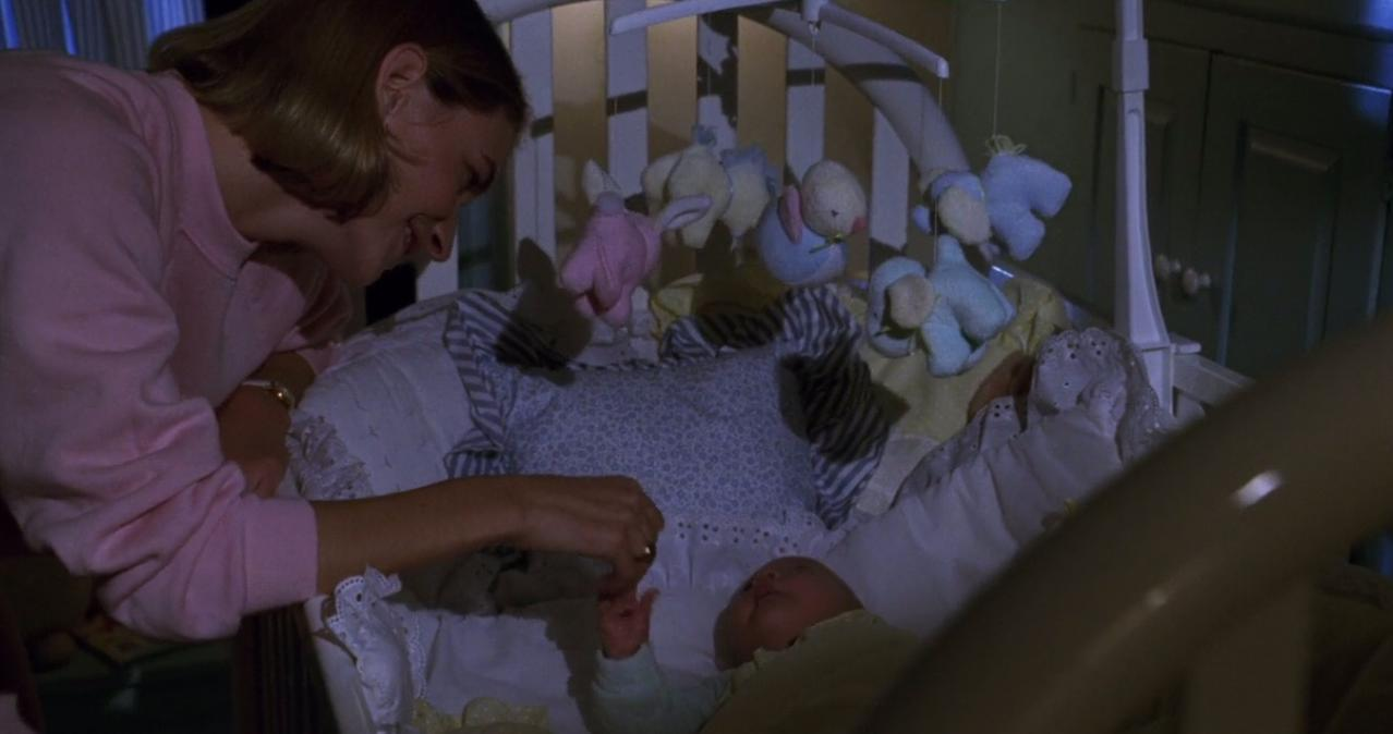 Eine Frau in einem rosa Pullover beugt sich über ein Baby in einem Kinderbett mit Spielzeug.