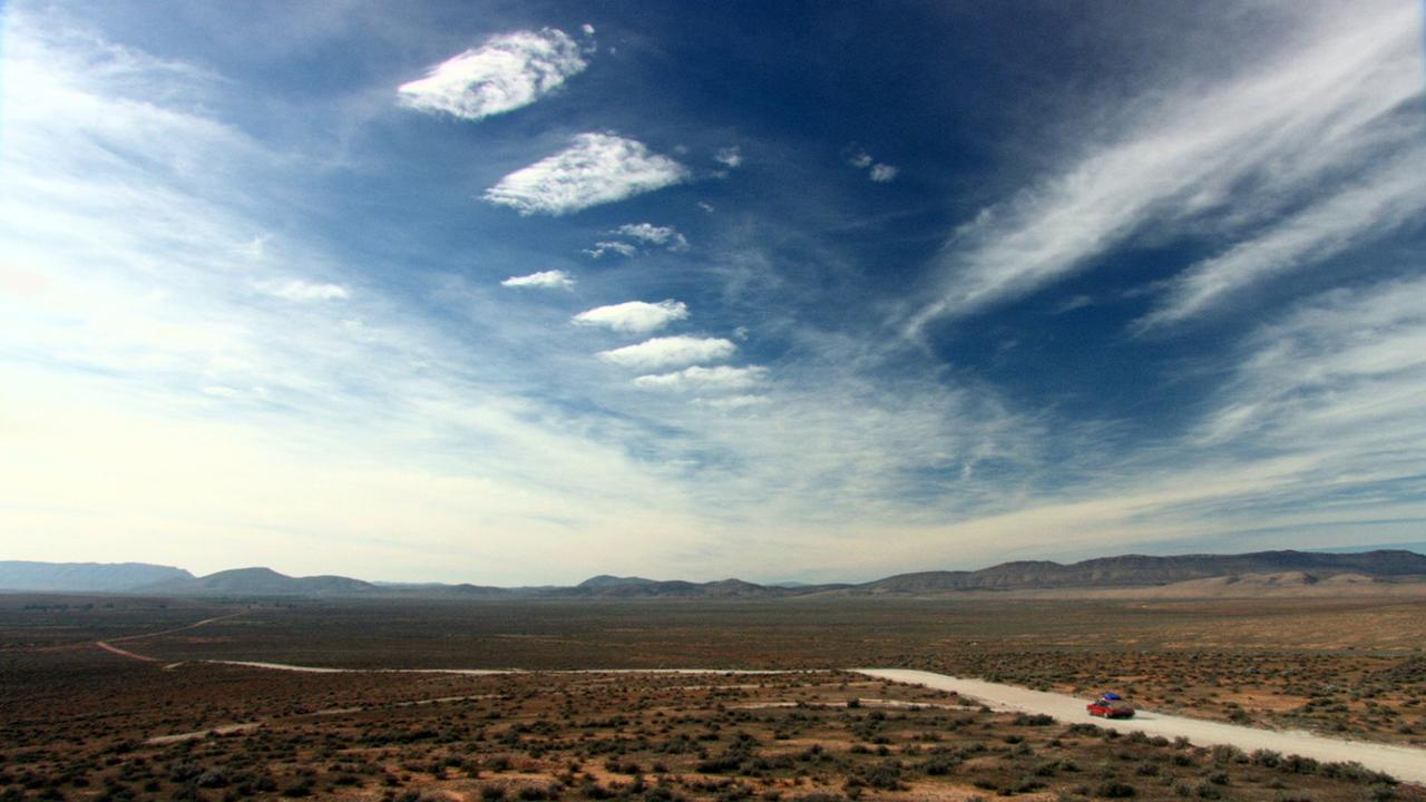 Ein rotes Auto fährt auf einer staubigen Straße durch eine weite, trockene Landschaft unter einem dramatischen Himmel.