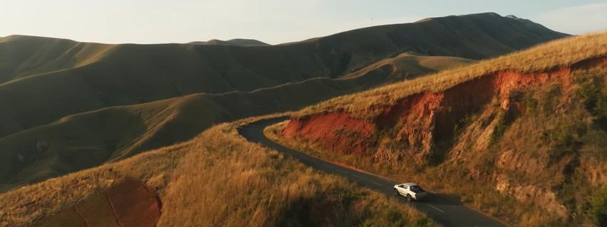 Ein weißes Auto fährt auf einer gewundenen Straße durch hügelige Landschaften.