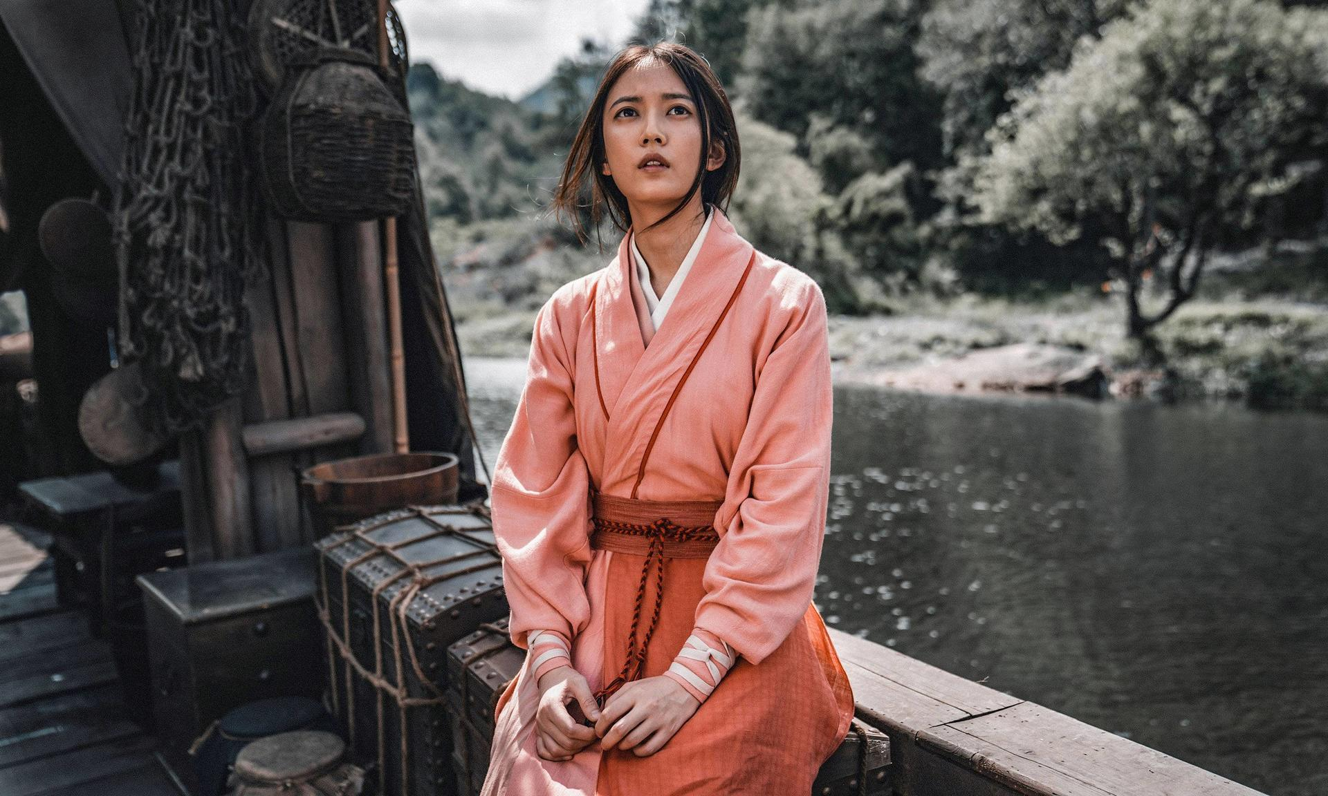 Eine junge Frau in einem rosa Kimono sitzt auf einem Boot, mit Wasser und Bäumen im Hintergrund.