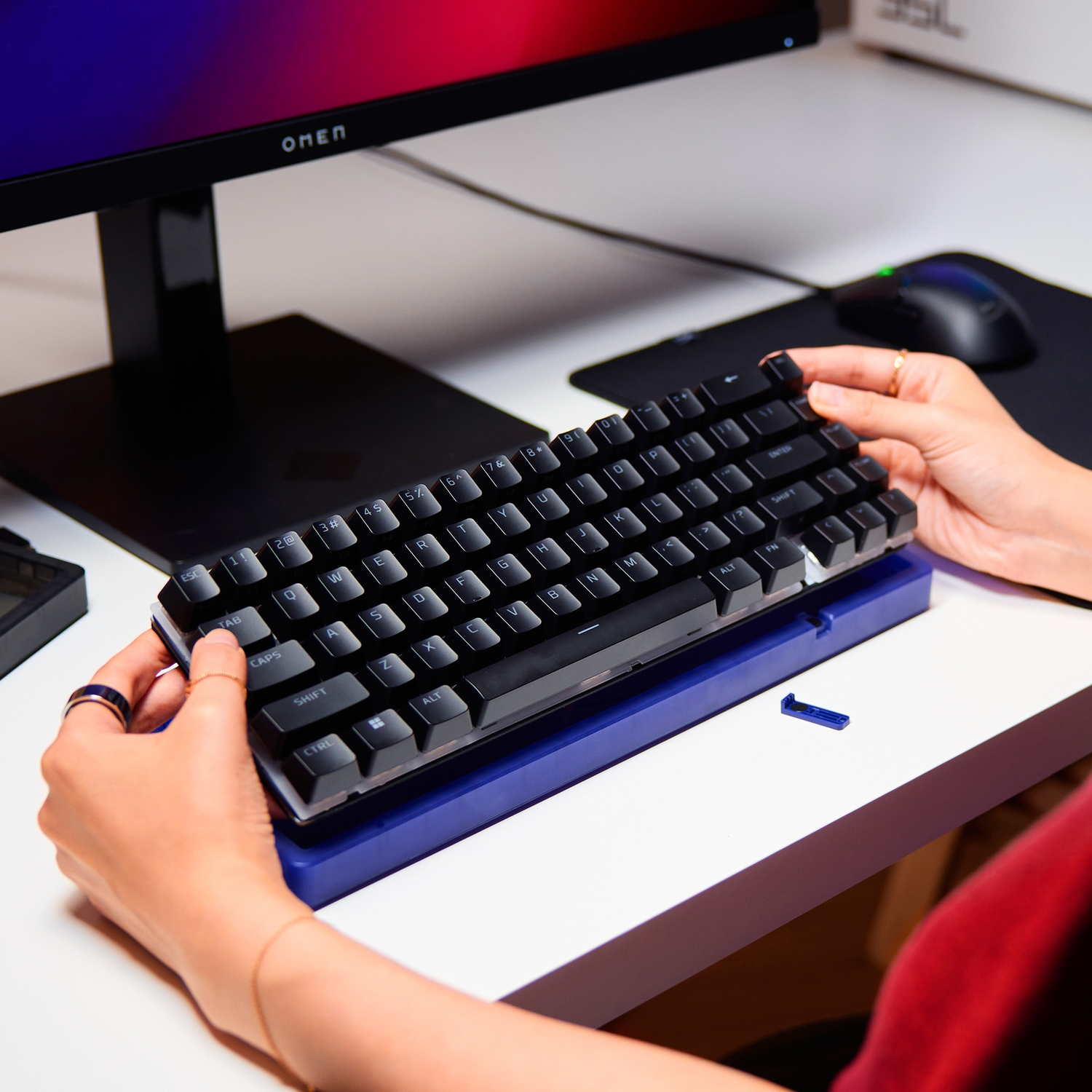 Hands adjust a black mechanical keyboard on a blue stand, with a monitor and mouse visible.