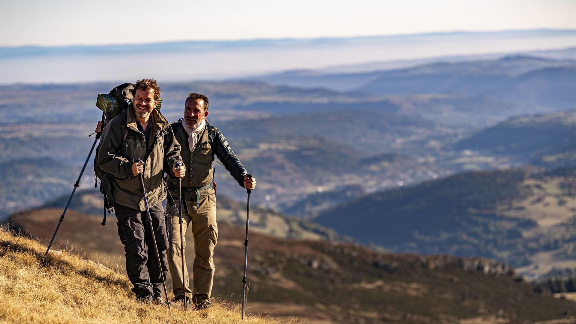 Zwei Männer wandern mit Stöcken auf einem grasbewachsenen Hügel mit Blick auf ein Tal.