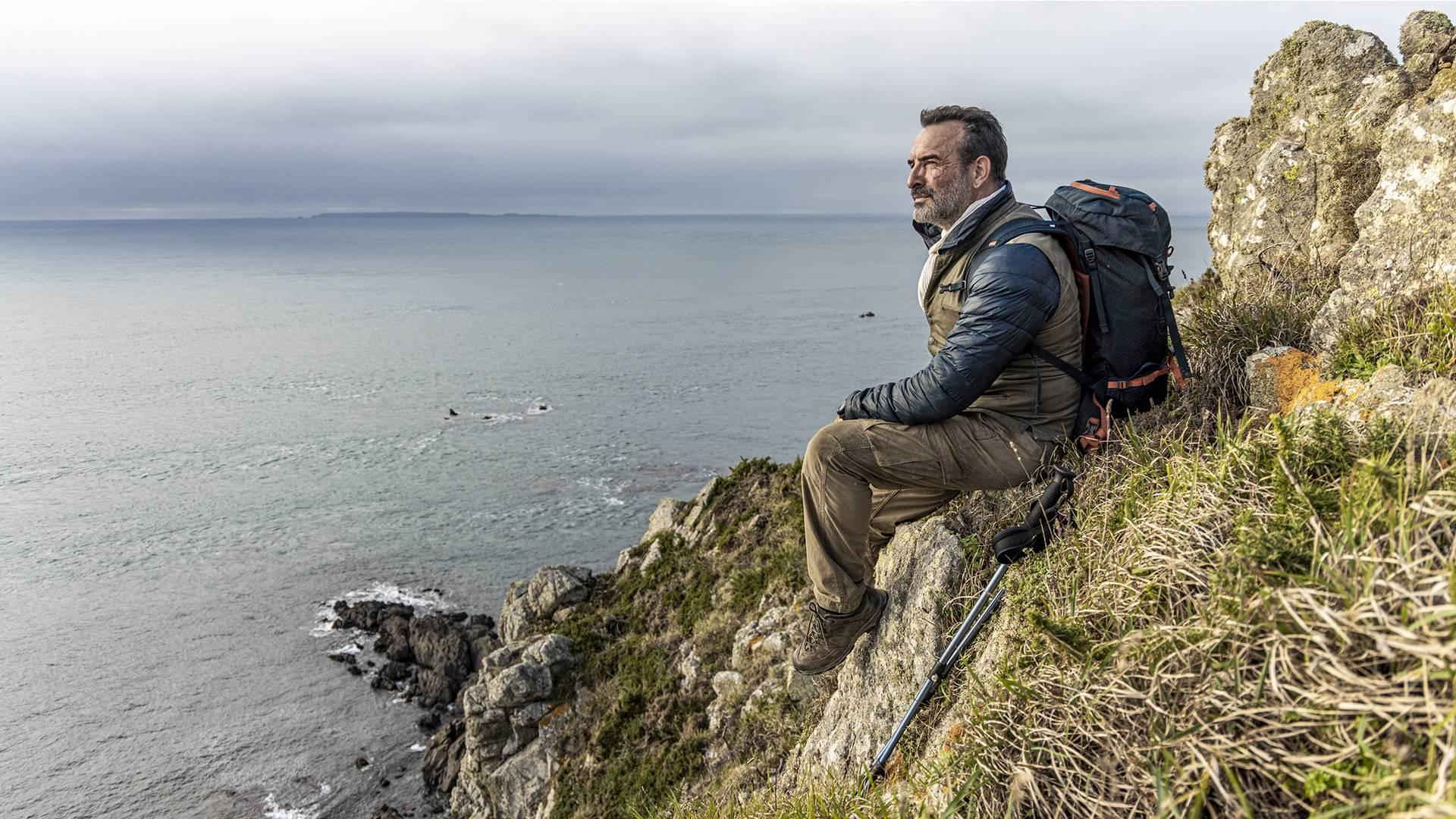 Ein Mann sitzt auf einer Klippe am Meer mit Rucksack und Wanderstöcken.
