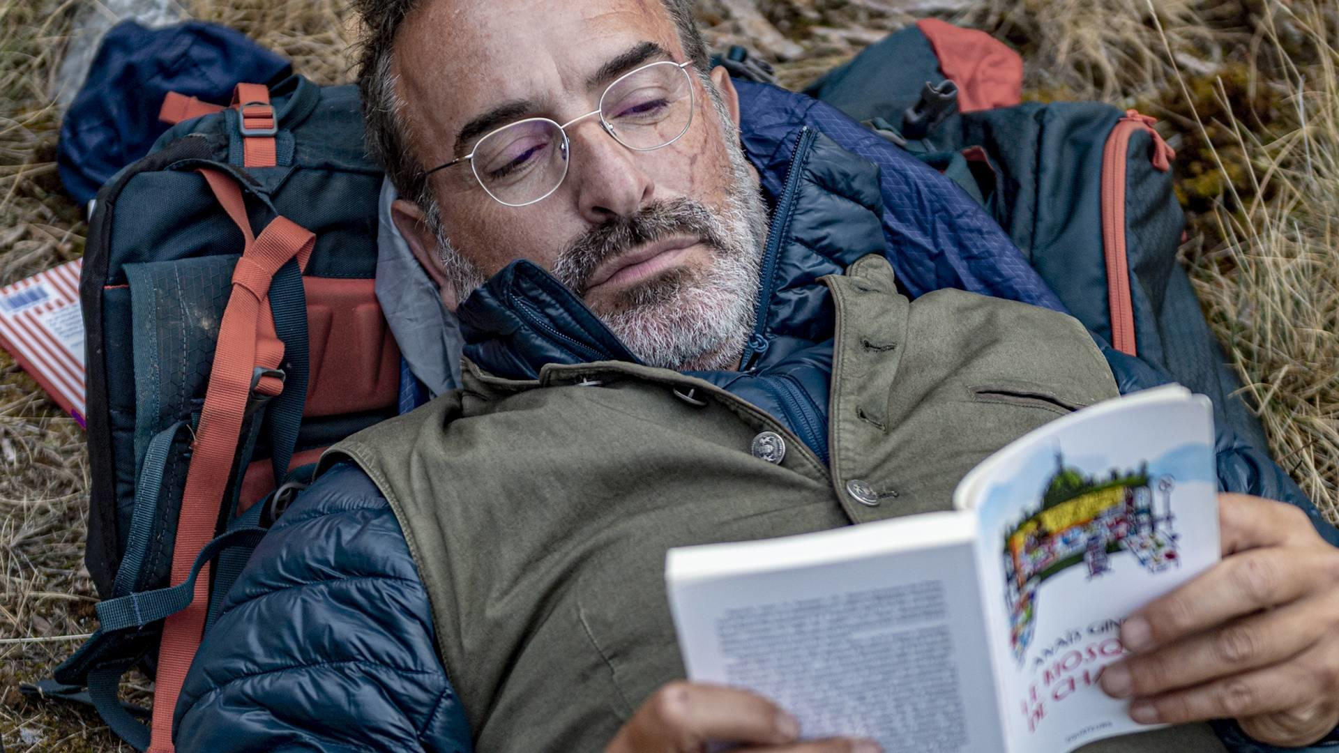 Mann liegt im Gras und liest Buch, Rucksack neben ihm.