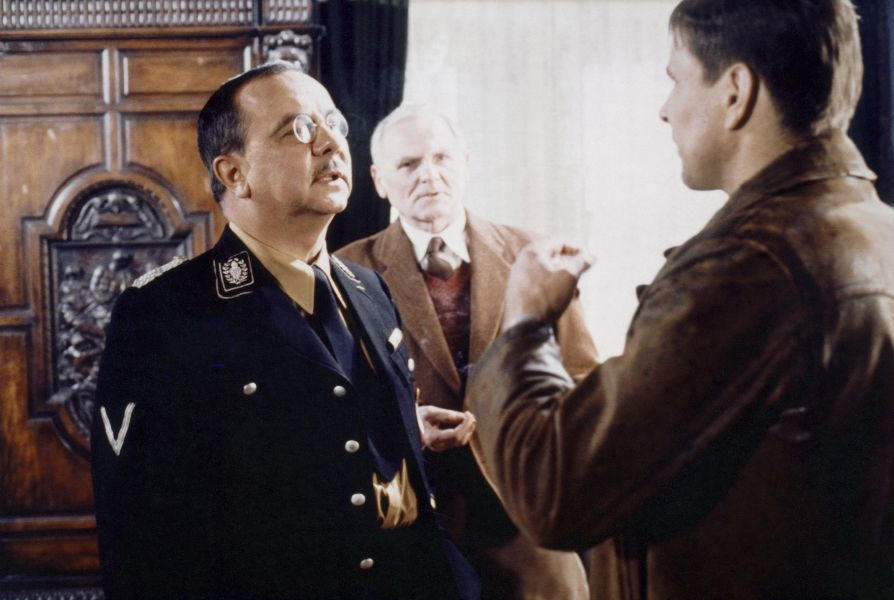 Ein Mann in Uniform spricht mit zwei Männern in einem Raum mit Holzakzenten.