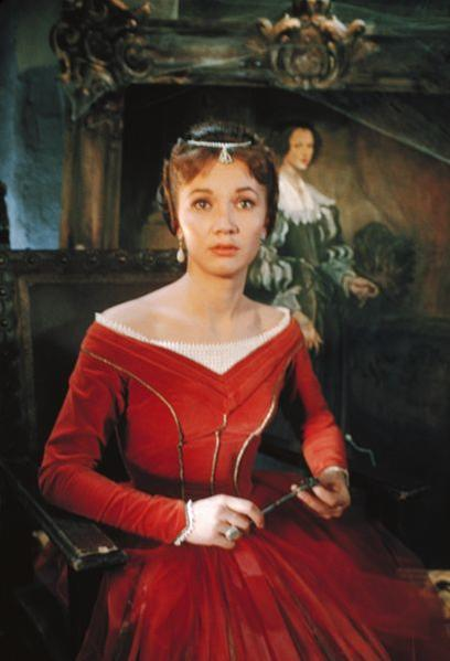 Eine Frau in einem roten Kleid mit Perlen-Diadem sitzt in einem dunklen Raum.