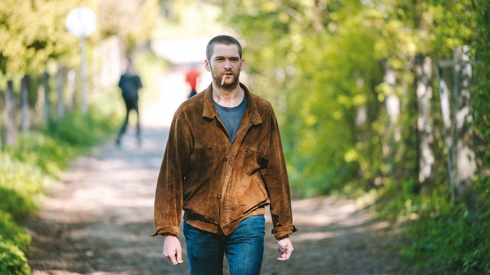 Ein Mann mit brauner Jacke und Zigarette geht auf einem Feldweg.