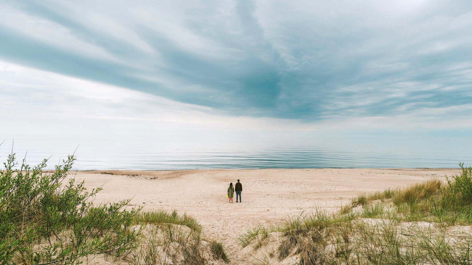 Zwei Personen stehen am Strand und blicken auf das Meer unter einem bewölkten Himmel.